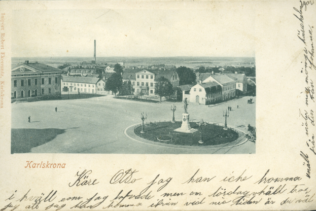 Alte Ansichtskarte zeigt eine Stadtansicht mit Gebäuden, Bäumen, Pfählen und einem zentralen Brunnen; handgeschriebener Text ist entlang der unteren Kante sichtbar.