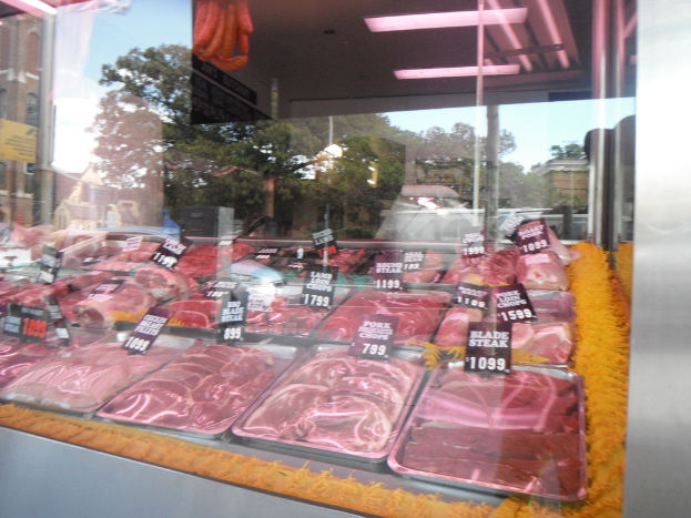 Eine Glasvitrine in einem Metzgerladen, die Tabletts mit Fleisch zeigt, mit Preisschildern, wobei durch das Fenster Gebäude und Bäume und die Decke des Raums oben im Bild zu sehen sind.