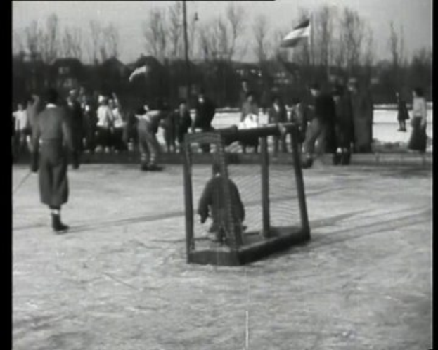 Schwarze und weiße Fotografie einer Gruppe von Menschen, die Hockey auf einem Eisplatz spielen, mit einem Netz in der Mitte, Bäumen, Pfählen und einer Fahne im Hintergrund und Text oben und unten.