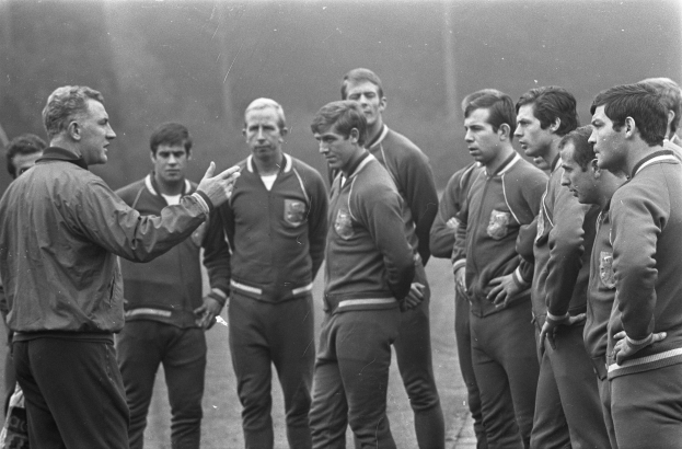 Schwarzes und weißes Foto einer Gruppe von Männern auf einem Fussballfeld mit der zentralen Figur in Wanderschuhen.