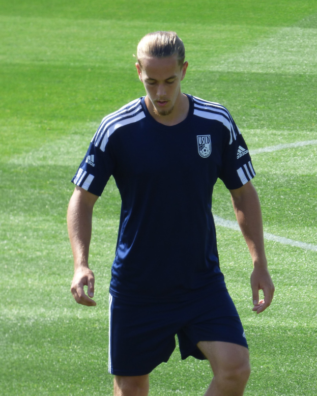 Ein professioneller Fußballspieler in einem blauen T-Shirt und Shorts geht selbstbewusst über ein saftiges grünes Feld mit einem entschlossenen Gesichtsausdruck.