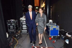 Angelina Jolie und John Kerry gehen über einen Boden, umgeben von Menschen, Geräten, Kabeln, einem Helm auf einem Wagen, einem Müllcontainer, einem Vorhang, einer Säule und einem Dach mit Deckenleuchten.