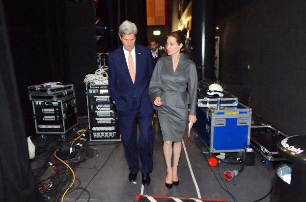 Angelina Jolie und John Kerry gehen über einen Boden, umgeben von Menschen, Geräten, Kabeln, einem Helm auf einem Wagen, einem Müllcontainer, einem Vorhang, einer Säule und einem Dach mit Deckenleuchten.