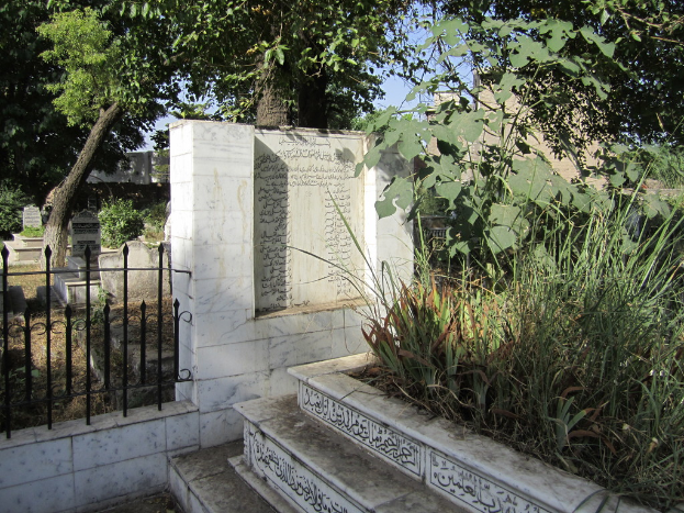 Ein Gedenkstein neben einem Geländer in einem Friedhof, mit Treppen unten, Gras und Bäumen rechts, Gräbern hinter dem Geländer und Gebäuden und Bäumen im Hintergrund.
