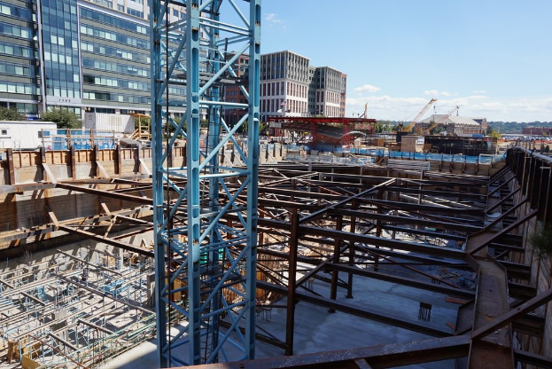 Das Bild zeigt eine Baustelle mit Gebäuden und Büros links, Eisenstangen und Maschinen rechts und diverser Ausrüstung und Kräne in der Mitte, unter einem Himmel im rechten oberen Eck.