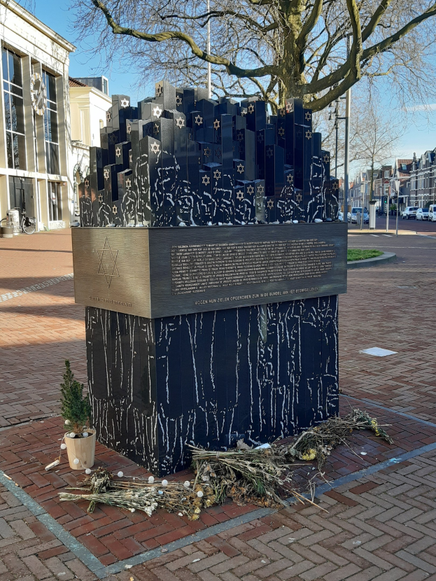 Denkmal in einem Stadtplatz gewidmet an Holocaust-Opfer, mit einem Baum davor, einem Topfgewächs, verstreuten trockenen Blättern und Gebäuden, Fahrzeugen, Pfählen und Bäumen im Hintergrund.