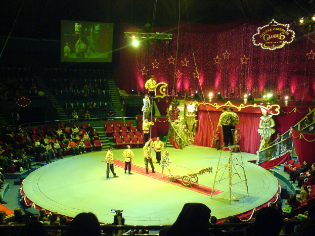 Eine Zirkusvorstellung mit Künstlern in der Mitte, Zuschauern auf der linken Seite, einem elektronischen Display oben und Lichtern und Puppen auf der rechten Seite.