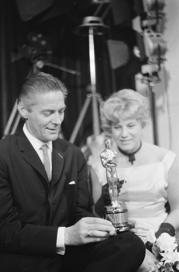 Ein Schwarz-Wei├č-Bild eines formell gekleideten Mannes, der eine Oscar-Statue hölt und eine Frau, die einen Blumenstrau├č hölt, nebeneinander sitzend mit Hintergrundbeleuchtung.