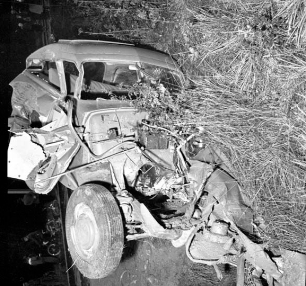 Ein Schwarz-Weiß-Foto eines schwer beschädigten Autos am Straßenrand mit einer eingedrückten Front, zersplitterten Fenstern, einem Baum im Hintergrund und verstreuten Gegenständen an der Szene.