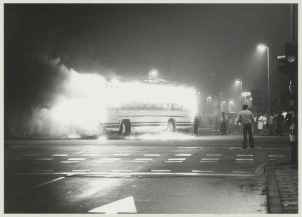 Ein Schwarz-Weiß-Foto eines brennenden Busses am Straßenrand mit einer Gruppe von Menschen auf dem Gehweg rechts und Straßenlaternen im Hintergrund.
