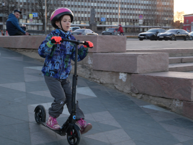 Ein junger Junge in Helm und Handschuhen fährt auf einem Roller über einen Gehweg, mit Treppen, Fahrzeugen, Menschen, Bäumen, Pfölen, Brettern, Gebäuden und einem klaren blauen Himmel im Hintergrund.