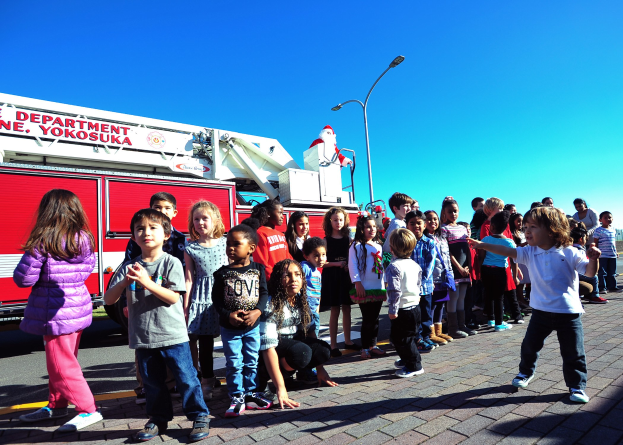 Eine Gruppe von Kindern vor einem Feuerwehrauto der Yokosuka-Feuerwehr während eines Weihnachtsumzugs, mit Laternenmasten, Bäumen und einem klaren blauen Himmel im Hintergrund.