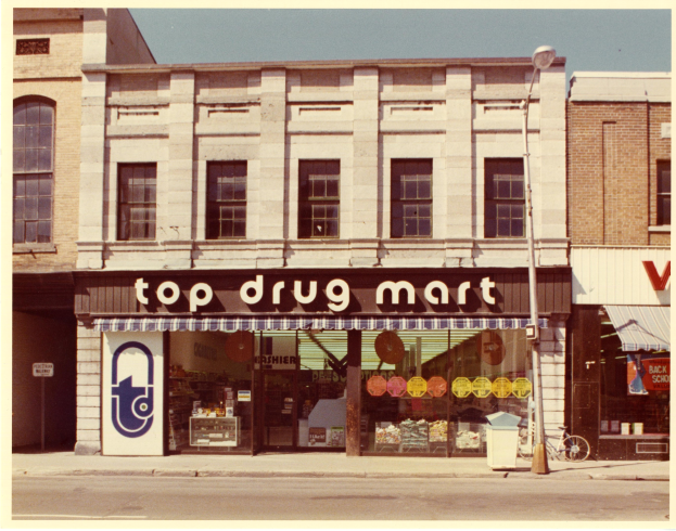 Straßensicht eines Top-Drogenladens mit einer Schildertafel, einem Fahrrad in der Nähe, einem Müllcontainer, einem Straßenpfosten und einem bewölkten Himmel.