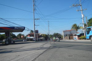 Ein blauer Lastwagen fährt auf einer Straße an einer Tankstelle vorbei, mit Strommasten und -leitungen entlang der Straße, Gebäuden, Bäumen und einem klaren blauen Himmel im Hintergrund.