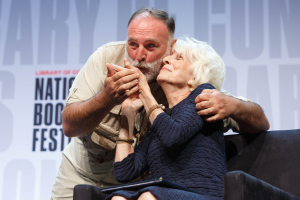 Ein Mann und eine Frau sitzen auf einer Couch und umarmen sich; die Frau trägt ein blaues Kleid, und ein weißes Banner mit "National Book Festival" ist im Hintergrund zu sehen.