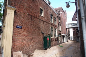 Ein Ziegelgebäude mit einem grünen Tor, umgeben von anderen Gebäuden mit Fenstern, einem Namensschild an der Wand, Rohren, Lampen, einer Brücke, Bäumen und anderen Gegenständen unter einem bewölkten Himmel.