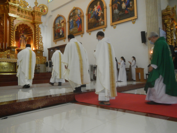 Eine Gruppe von Priestern geht einen roten Teppich in einer Kirche entlang, während Menschen auf dem Boden vor und rechts von ihnen stehen und Foto Rahmen an der Hintergrundwand hängen.