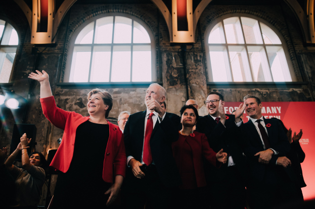 Eine Gruppe von Menschen, wahrscheinlich Labour-Parteimitglieder, steht klatschend vor einer Menge mit einem Banner, Glasfenstern und beleuchteten Lichtern hinter ihnen, feiert ihren Wahlsieg von 2019.