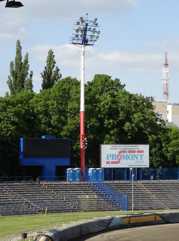 Rennstrecke umgeben von Gras, Geländern, Stufen, Leuchtenmasten, einem Schild, Bäumen, Gebäuden, Türmen und einem bewölkten Himmel.