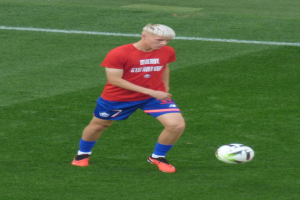 Ein junger Mann in rotem T-Shirt und blauen Shorts, der auf einem grünen Rasen Fußball spielt und Fußballschuhe trägt.