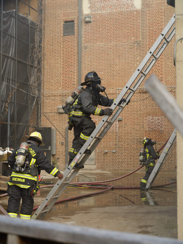 Feuerwehrleute in Helmen und Ausrüstung, die eine Leiter vor einem Backsteingebäude hinaufsteigen, mit Rohren auf dem Boden und einer Metallstange am unteren Ende, mit einem weiteren Gebäude mit Fenstern und einem Netz im Hintergrund.