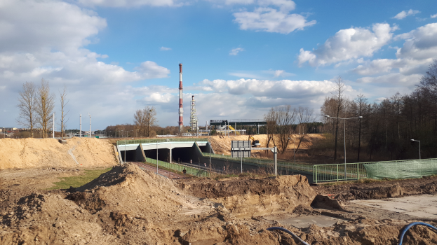 Eine Baustelle mit einem großen Haufen Erde im Vordergrund, umgeben von Bäumen, Pfählen, Lampen, Geländern und einer Brücke, mit Türmen und einem bewölkten Himmel im Hintergrund und Erde und Rohren unten.