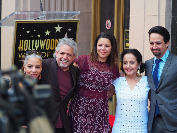 Eine Gruppe von Menschen steht vor einem Stern auf dem Hollywood Walk of Fame, mit einem Rednerpult und Mikrofon in der Mitte, einer Person mit einer Kamera links daneben und einer Tafel mit Text dahinter, mit Wänden und einer Tür im Hintergrund.