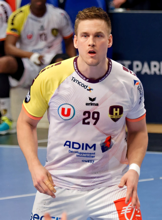 Ein Mann in weißem Hemd und gelben Shorts spielt Handball, hält ein Objekt und hat einen entschlossenen Gesichtsausdruck, vor einer Tafel mit der Aufschrift "Ligue 1 - Futsal - Olympique de Marseille" und ein paar im Hintergrund sitzenden Menschen.