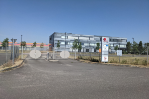 Großes industrielles Gebäude mit der Aufschrift "First Solar" und vielen Fenstern, umgeben von einem Metallzaun, umgeben von Bäumen, Pflanzen, Gras, Schildern, Straßenlaternen und einem Weg unter einem bewölkten Himmel.