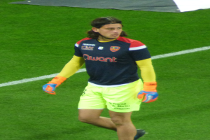 Ein Mann in einem blauen T-Shirt und gelben Shorts läuft auf einem Fußballfeld mit ausgestreckten Armen und entschlossener Miene, mit einem Tuch oben auf dem Bild.