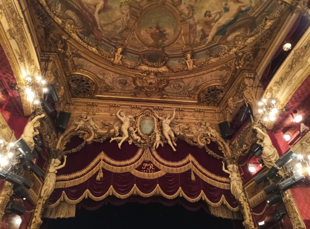 Theaterinnenraum mit rotem Vorhang, Kronleuchtern, Skulpturen, Lampen und einer Deckenmalerei, mit einer Bühne mit ein paar Menschen davor.