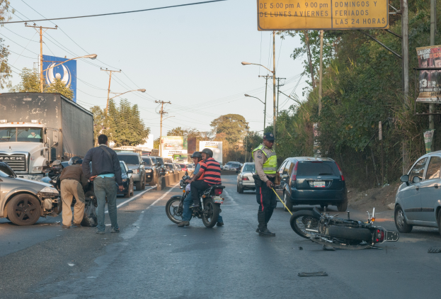 Eine Gruppe von Menschen umringt ein verunglücktes Motorrad am Straßenrand mit mehreren Fahrzeugen, darunter ein Lastwagen, und einer Hintergrundlandschaft aus Bäumen, Pfählen, Lampen und Schildern unter einem Himmel.