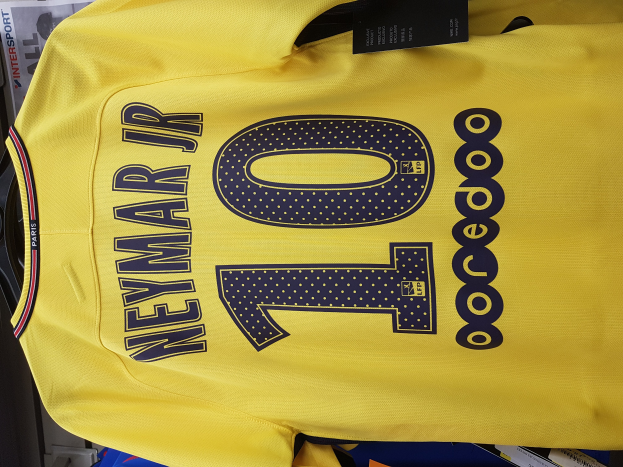 Ein gelbes Fußballtrikot mit der Nummer 10, das Neymar Jr. von der Heimkollektion des PSG 2018-19 gehört, wird mit einem schwarzen Etikett und Papieren sowie anderen Gegenständen im Hintergrund gezeigt.