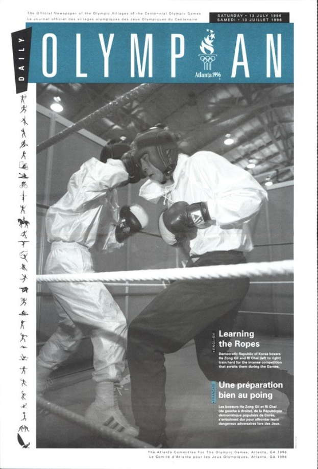 Schwarzes und weißes Foto von zwei Boxern in einem Ring mit Helmen und Handschuhen, umgeben von Seilen und beleuchtet von Scheinwerfern, mit 'Olympian Magazine'-Text oben und unten.