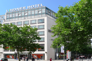 Großes modernes Hotelgebäude mit Glasfenstern und Beschriftung oben, umgeben von Bäumen und einer belebten Straße mit Fußgängern und Fahrzeugen unter einem sichtbaren Himmel.