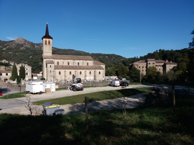 Luftaufnahme einer kleinen Stadt mit einer zentralen Kirche, umgeben von Gebäuden, Bäumen, Fahrzeugen, Straßen und Schildern, vor einem Hügel- und einem klaren blauen Himmel.