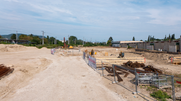 Eine Baustelle mit Erde, Metallstangen, Holzobjekten, einem Zaun, einer Fahne mit Text, Bäumen, Pfählen mit Drähten, Fahrzeugen, Gebäuden mit Fenstern, Hügeln und einer bewölkten Himmel.
