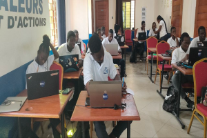 Eine Gruppe von Menschen sitzt an Schreibtischen mit Laptops in einem Klassenzimmer, mit Taschen auf dem Boden und Text an der linken Wand, was auf einen Online-Kurs in Ghana hinweist.