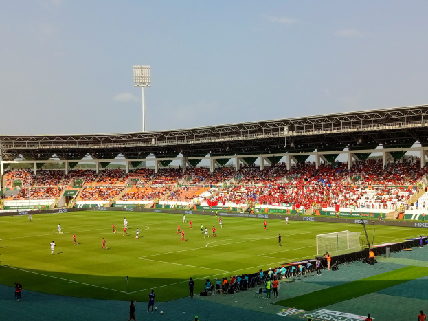 Ein Fussballspiel wird in einem grossen Stadion gespielt, mit Spielern auf dem Feld, einem Tor, einem Zaun, einer Fahne mit Text, Metallrahmen, einem Dach mit Deckenleuchten und einem bewölkten Himmel.