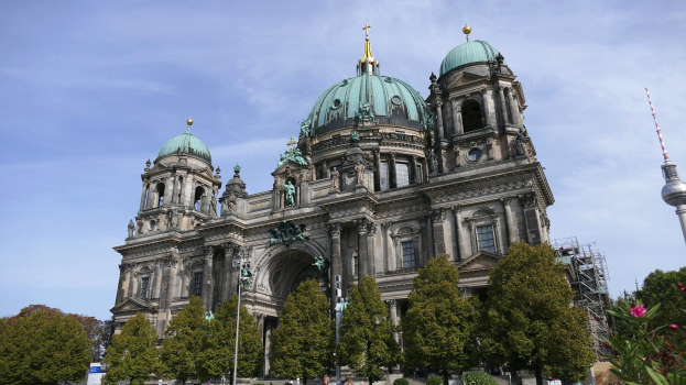 Der Berliner Dom, eine große Kathedrale mit Fenstern, Säulen, Bögen und Statuen, steht prominent in Berlin, Deutschland, mit Menschen davor, Bäumen, blühenden Pflanzen und einem Turm in der Nähe unter einem bewölkten Himmel.