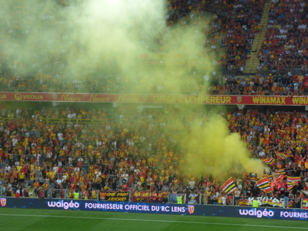 Ein Fußballstadion voller Menschen, einige halten Fahnen und Schilder, andere stehen oder sitzen, mit einer Wolke aus Rauch in der Luft.