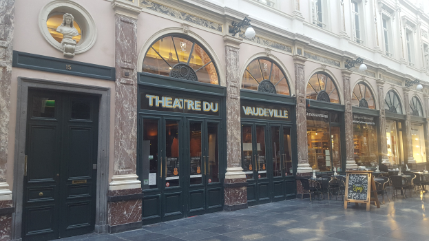 Theater du Vaudeville-Gebäude in Paris mit Türen, Fenstern, Lichtern, Texttafeln, Tischen, Stühlen und einer Wandstatue.