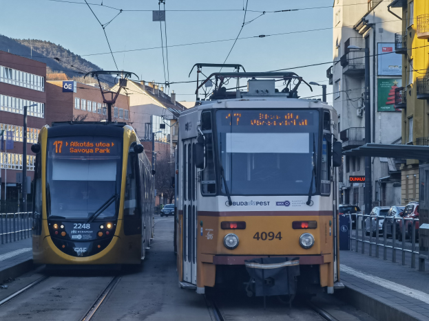 Zwei gelbe und weiße Straßenbahnen fahren auf Stadtbahngleisen neben hohen Gebäuden, mit Fahrzeugen auf der Straße, Geländern auf beiden Seiten und einem Hintergrund mit Gebäuden, Pfählen, Schildern, Bäumen, einem Hügel und dem Himmel.