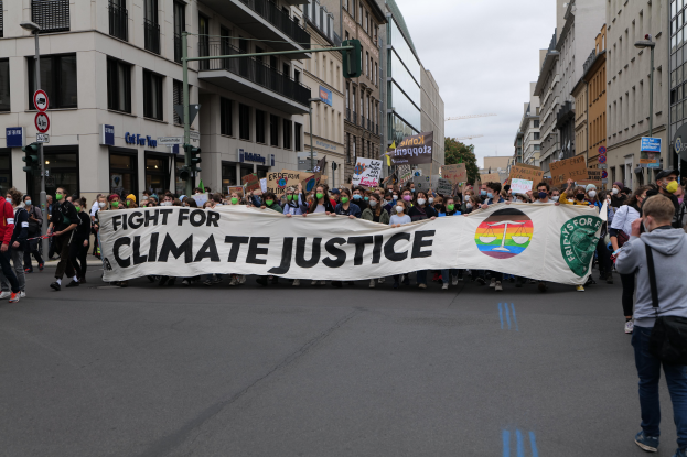 Eine Gruppe von Menschen marschiert mit einem "Kampf für Klimagerechtigkeit"-Schild die Straße entlang, mit Gebäuden, Schildern, Ampeln und Bäumen an der Straße unter einem bewölkten Himmel.