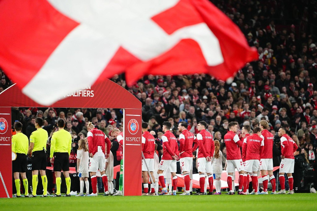 Eine Gruppe von Menschen steht auf einem Fussballfeld mit einer roten und weissen Flagge im Vordergrund und einem Bogen im Hintergrund, auf dem der Text 'Bayern Munich vs Bayern Munich Betting Tips & Preview' angezeigt wird, mit einer grossen Menschenmenge im Stadion.