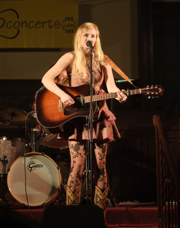 Eine Frau mit Gitarre singend auf der Bühne bei einem Konzert, im Hintergrund eine Band und an der Wand ein Bildschirm.