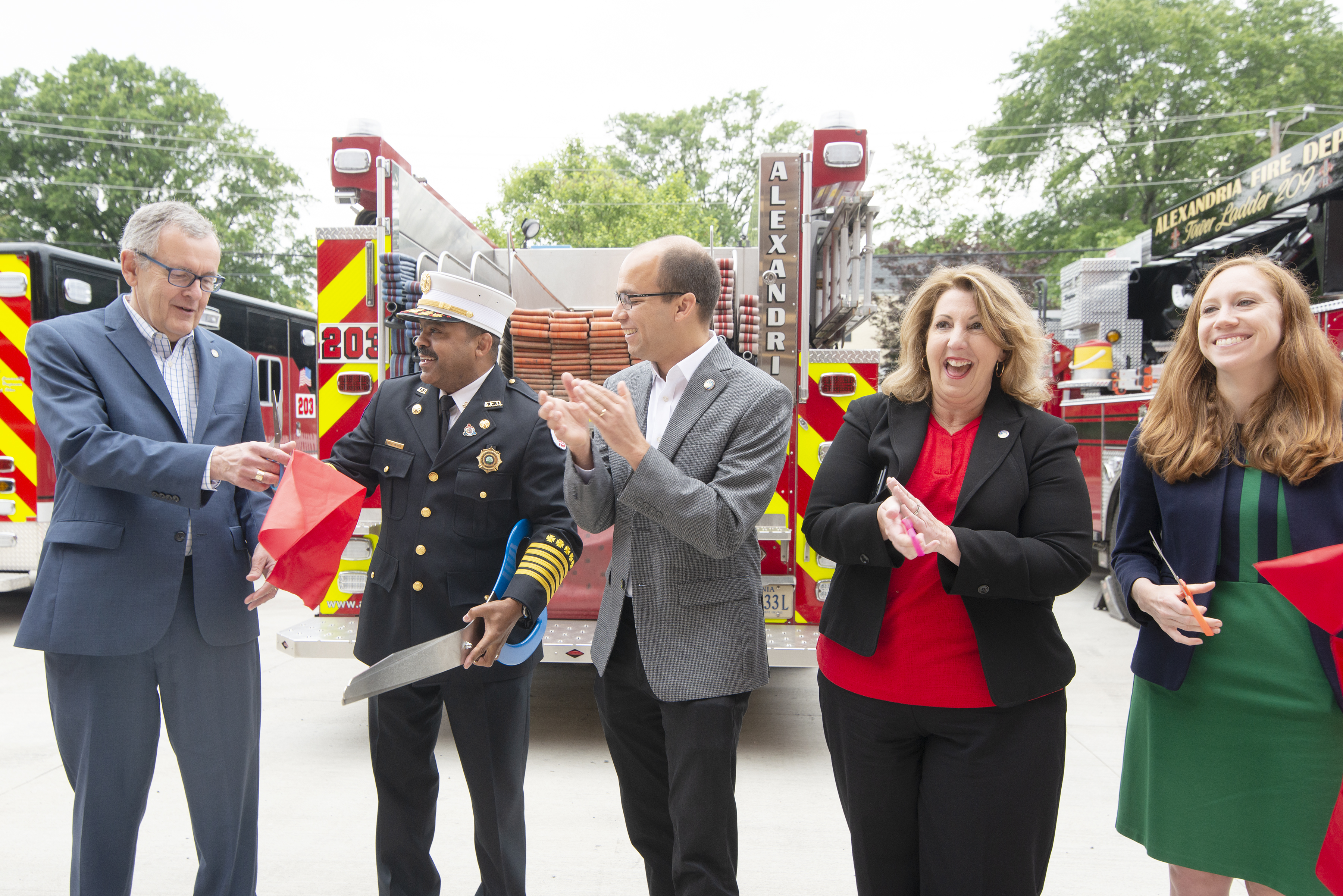 Eine Gruppe von Menschen steht vor einem Feuerwehrauto, klatscht und lächelt, während zwei eine Schere und ein rotes Tuch halten, bei einer Einweihungszeremonie für die Alexandria Fire Department, mit Bäumen, Drähten und einem klaren blauen Himmel im Hintergrund.