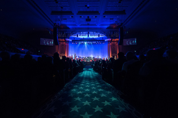 Ein großer Saal mit Sitzplätzen, eine Bühne mit Musikern und Deckenbeleuchtung und -fernseher, was auf einen Konzerthallensaal während der Nationalkonvention 2019 hinweist.