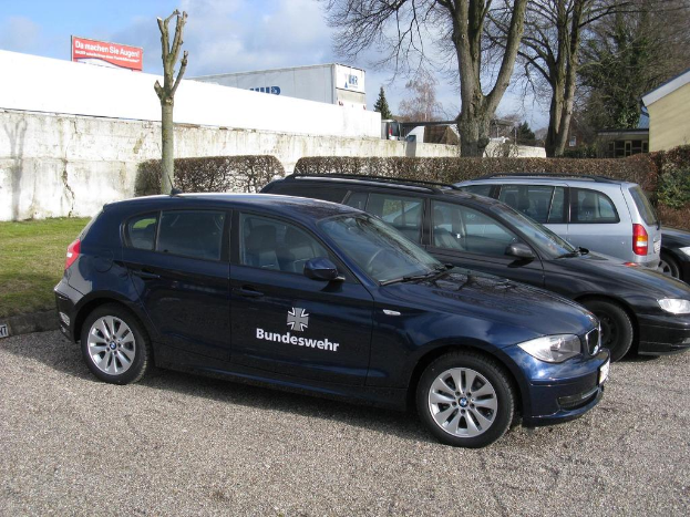 Mehrere Autos auf einem Weg umgeben von grünem Gras, mit Büschen, Bäumen und Gebäuden im Hintergrund, ein Schild mit Text und ein Logo sichtbar.