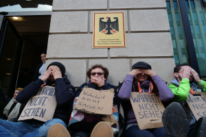 Eine Gruppe von Menschen sitzt vor einem Gebäude, hält Schilder hoch, einige decken ihre Gesichter ab, mit einem Logo und einer Tafel an der Wand und einer Glastür auf der rechten Seite.